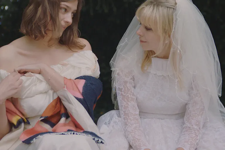 Two women sitting and looking at each other. One is wearing a wedding dress and the other one a formal white dress and a colourful shawl