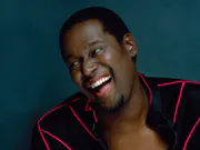A Black Man in a black jacket with bright red lines is laughing and leaning to his right
