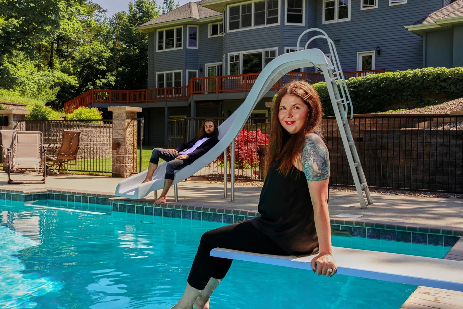 A woman sitting on a pool platform smiles looking at the camera. Further back a man sits on a slide by the same pool with a serious look facing the camera
