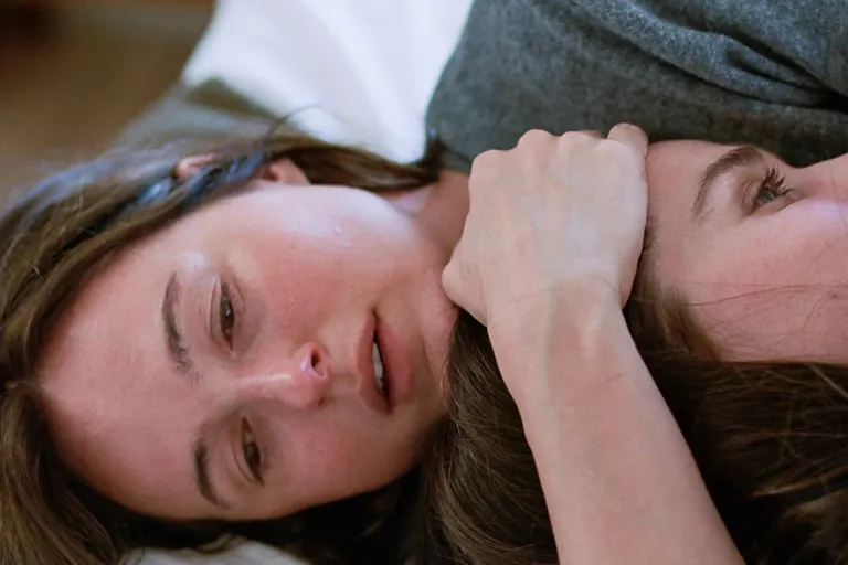A close up of a woman, lying down, holding another woman’s head to her chest.