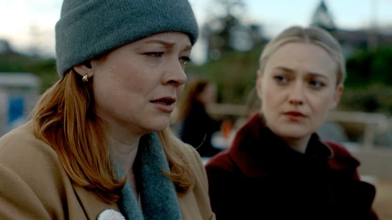A close-up of Marissa Irvine, played by Sarah Snook, and Jenny Kaminski, played by Dakota Fanning. They are outside. Marissa is wearing a grey hat, scarf and a camel coat. She looks upset. Jenny, wearing a claret coloured coat looks sympathetically at Marissa.