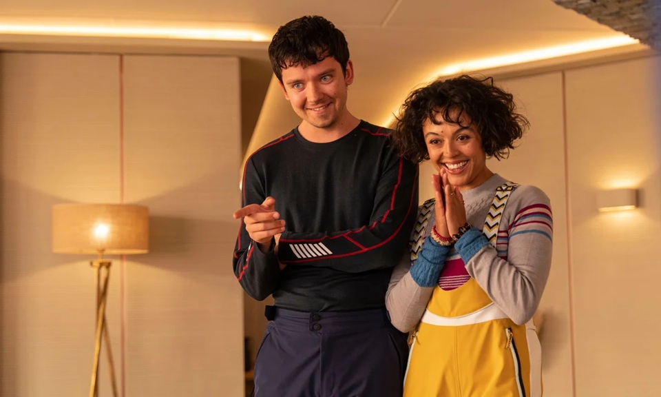 A young white man dressed all in dark ski clothes, stands in a warmly lit room beside a young Black woman in bright, colourful ski clothes, both smiling