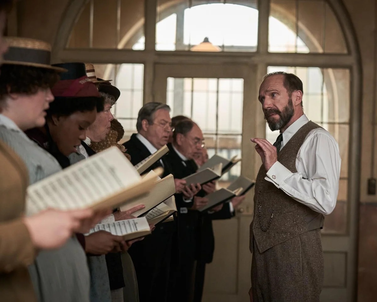 Ralph Fiennes, playing a choir master, stands, in profile, in front of a line of people holding choir books. The people are singing. Everyone is dressed in early twentieth century clothing.
