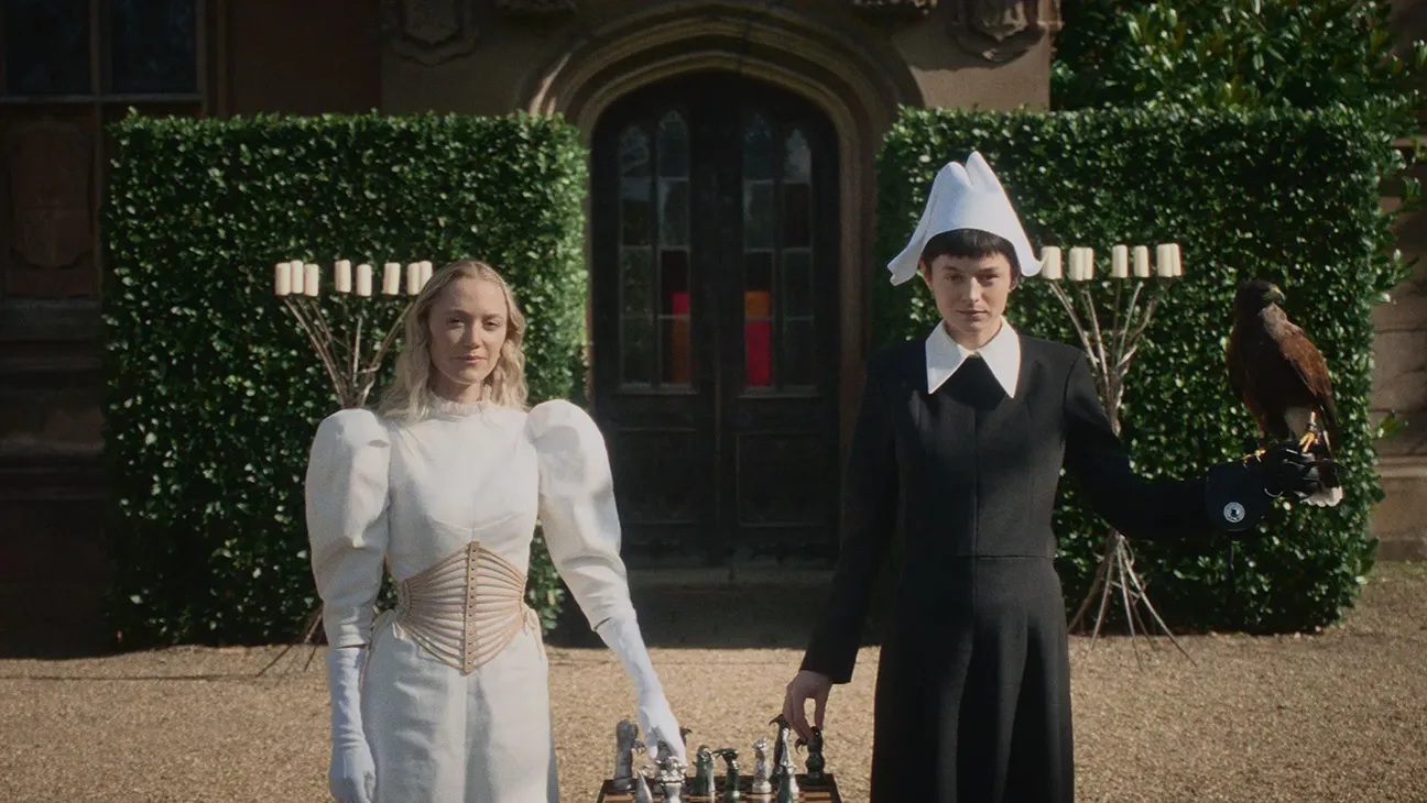 Two women stand outside facing us, behind them a tall hedge and an old door to what looks like an old country mansion. It is sunny. One woman wears white, the other black and between them is a chess board. Each woman holds a piece on the board from an outstretched arm. On the other arm of the woman in black is a hawk.