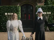 Two women stand outside facing us, behind them a tall hedge and an old door to what looks like an old country mansion. It is sunny. One woman wears white, the other black and between them is a chess board. Each woman holds a piece on the board from an outstretched arm. On the other arm of the woman in black is a hawk.