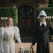 Two women stand outside facing us, behind them a tall hedge and an old door to what looks like an old country mansion. It is sunny. One woman wears white, the other black and between them is a chess board. Each woman holds a piece on the board from an outstretched arm. On the other arm of the woman in black is a hawk.