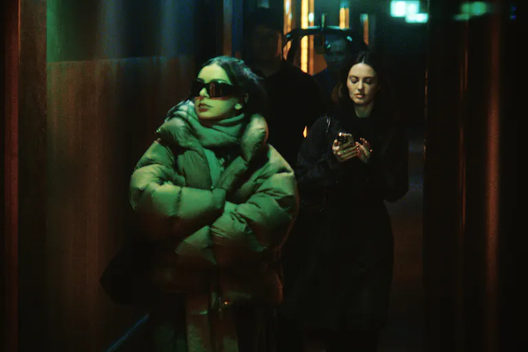 A young woman in oversized glasses and puffer coat walk with an entourage behind her
