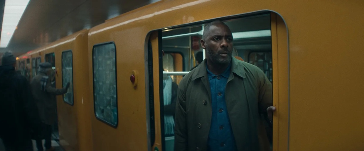 A Black man in a mac stands in between an open door of a bright yellow train