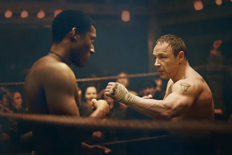 A young Black man and an older white man circle each other in a boxing ring
