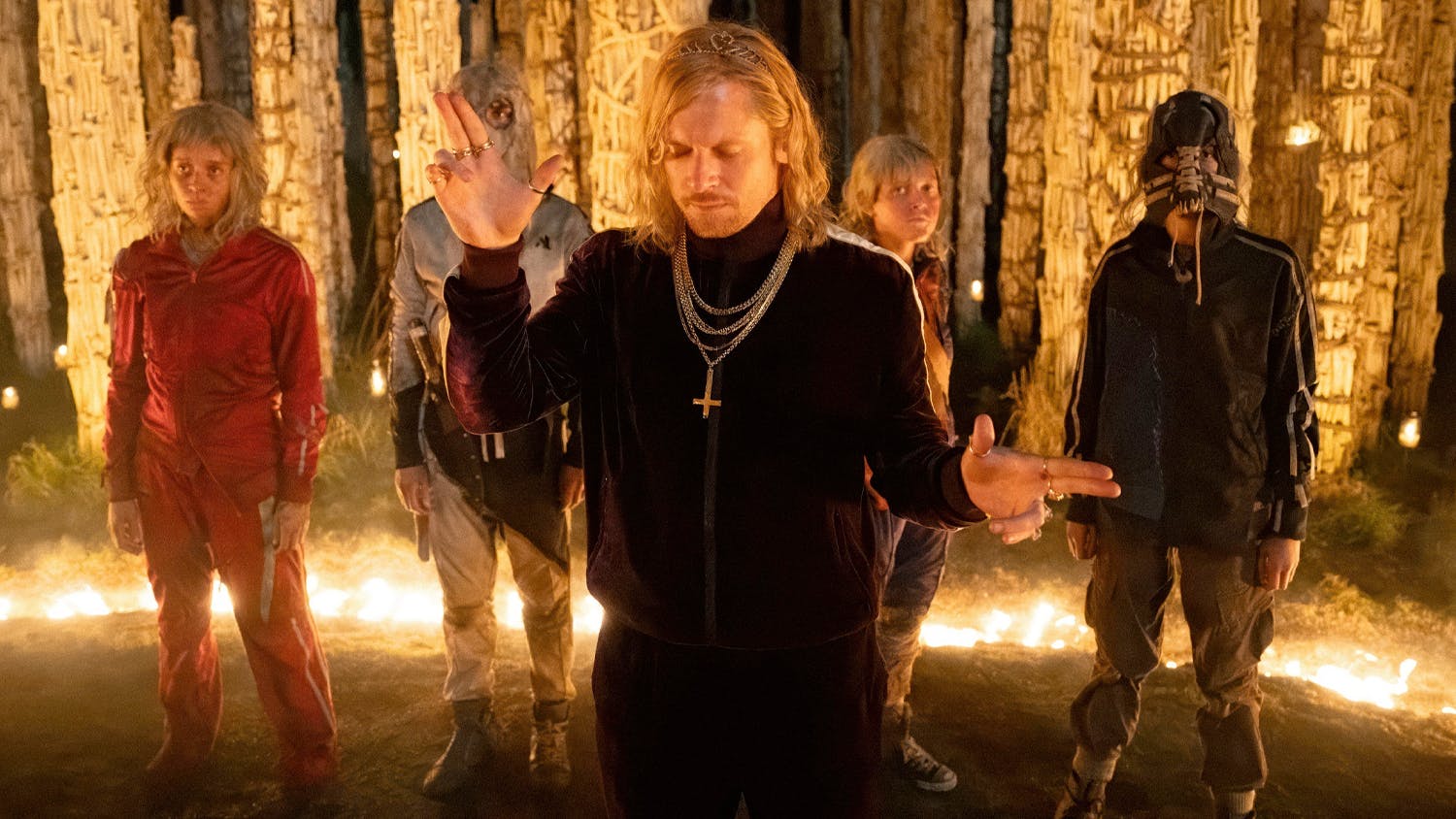 Five young people in tracksuits and gold chains standing in front of lit up pillars of bones and skulls, a long haired blond man at the lead