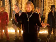 Five young people in tracksuits and gold chains standing in front of lit up pillars of bones and skulls, a long haired blond man at the lead
