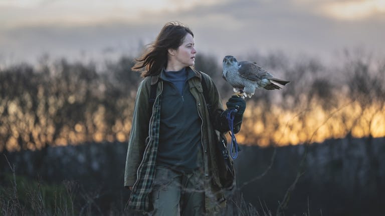 A white woman in a cold field at dusk looking at a hawk as it's perched on her arm
