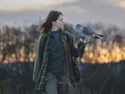 A white woman in a cold field at dusk looking at a hawk as it's perched on her arm
