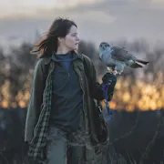 A white woman in a cold field at dusk looking at a hawk as it's perched on her arm