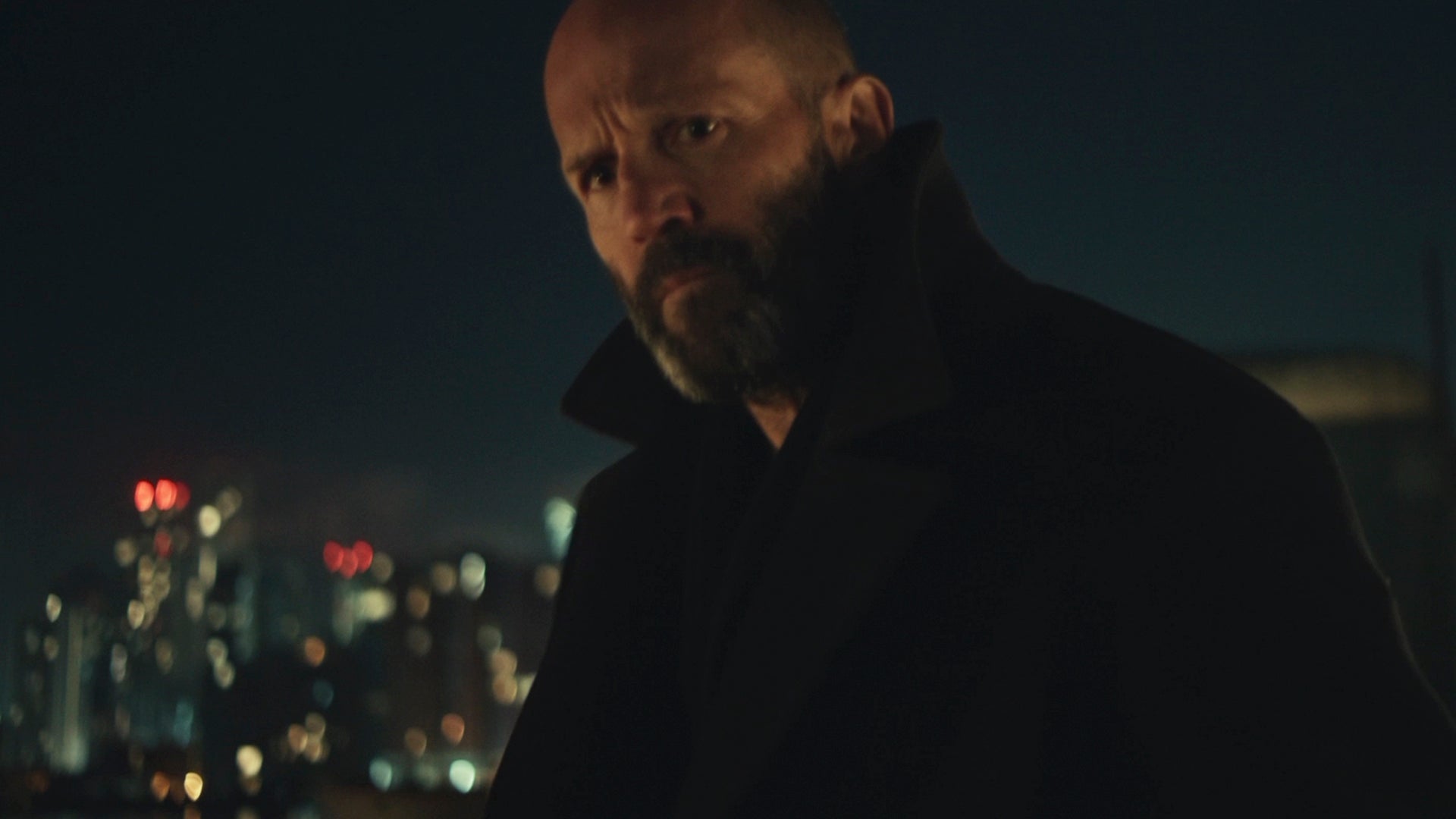 A white man with a short beard wearing a black wool coat with the collar turned up looking menacing with a night cityscape behind him