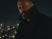 A white man with a short beard wearing a black wool coat with the collar turned up looking menacing with a night cityscape behind him