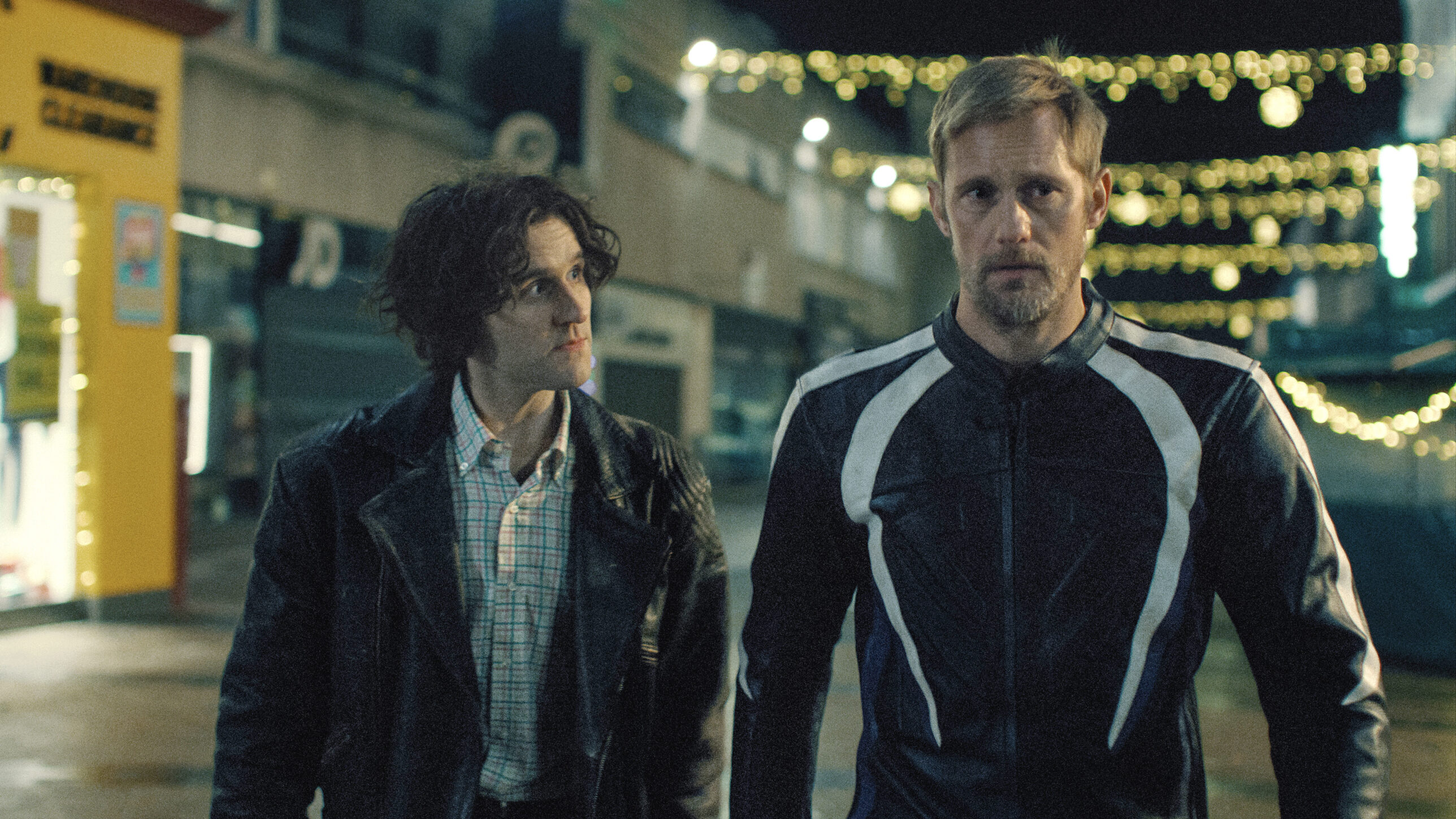 A young white man with floppy hair looks intently at a tall older white man in bike leathers