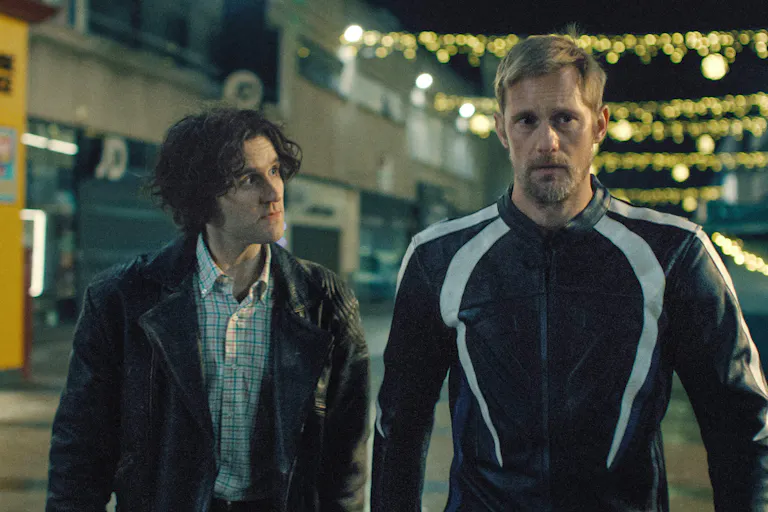 A young white man with floppy hair looks intently at a tall older white man in bike leathers