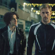 A young white man with floppy hair looks intently at a tall older white man in bike leathers