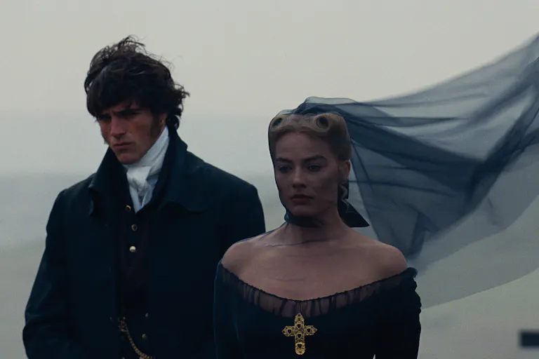 A young white man and woman in black 18th century clothing, her black veil whipping in the wind as they stand on the Yorkshire moor looking somber