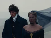 A young white man and woman in black 18th century clothing, her black veil whipping in the wind as they stand on the Yorkshire moor looking somber