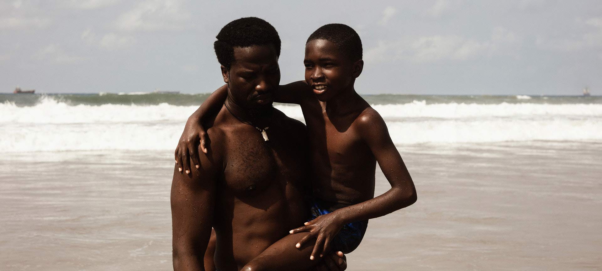 A Black man holding a young Black boy in his arm as they stand on a beach
