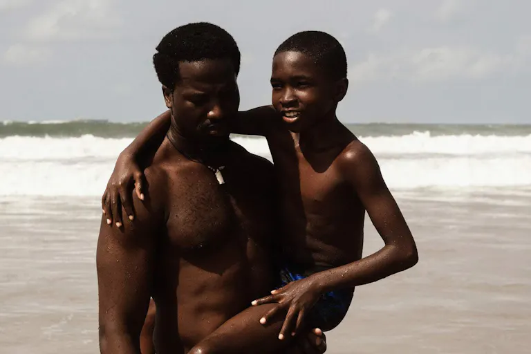 A Black man holding a young Black boy in his arm as they stand on a beach