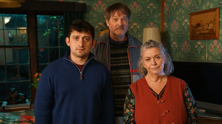 A young white man, older man and woman stand in a living room looking concerned