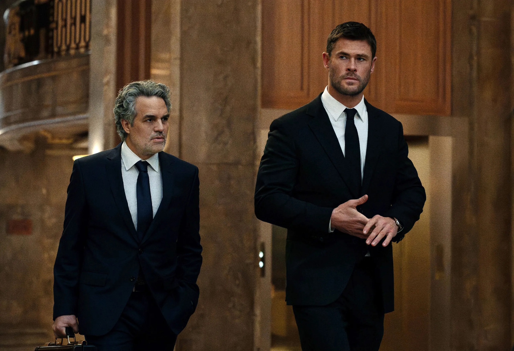 Two men in sharp suits walk through a marble foyer