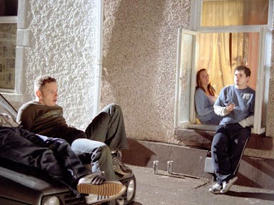 Three teen boys and one teen girl lounge on the bonnet of a car and sit outside a windowsill smoking and drinking cans