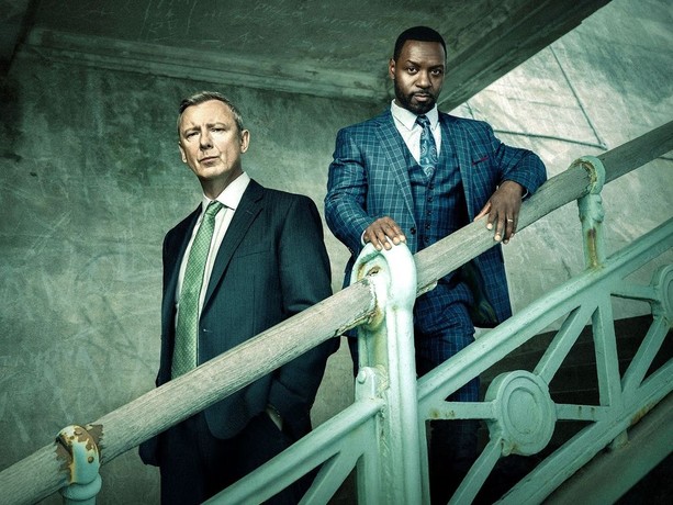 A white man and a black man stand on a staircase in suits looking directly at the camera.