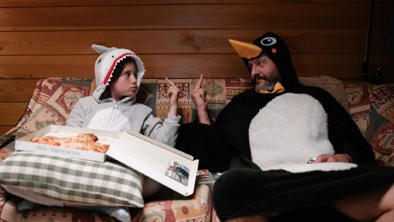 A boy (left) sits with a pizza box in his lap in a shark onesie, a man (right) sits in a penguin onesie. Both are holding up their middle finger at the other.