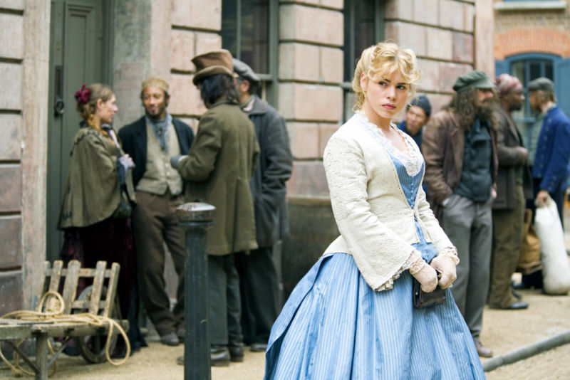 A blonde, white woman stands in a blue dress with a white petticoat looking into the distance with suspicious eyes.
