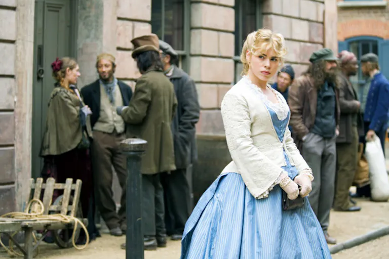 A blonde, white woman stands in a blue dress with a white petticoat looking into the distance with suspicious eyes.