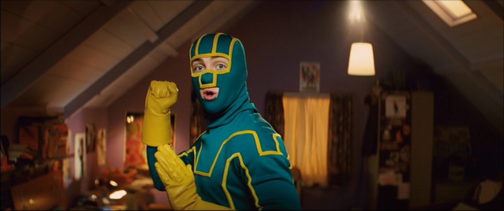 A white man stands in a fighting position in a teal and yellow superhero costume with his face covered except his mouth and eyes.