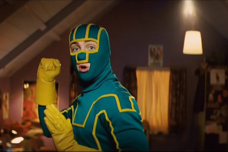 A white man stands in a fighting position in a teal and yellow superhero costume with his face covered except his mouth and eyes.