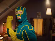 A white man stands in a fighting position in a teal and yellow superhero costume with his face covered except his mouth and eyes.