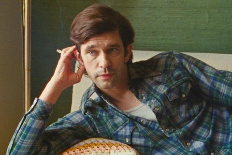 A white man lies horizontally in a blue and green plaid shirt with a cigarette in his hand.