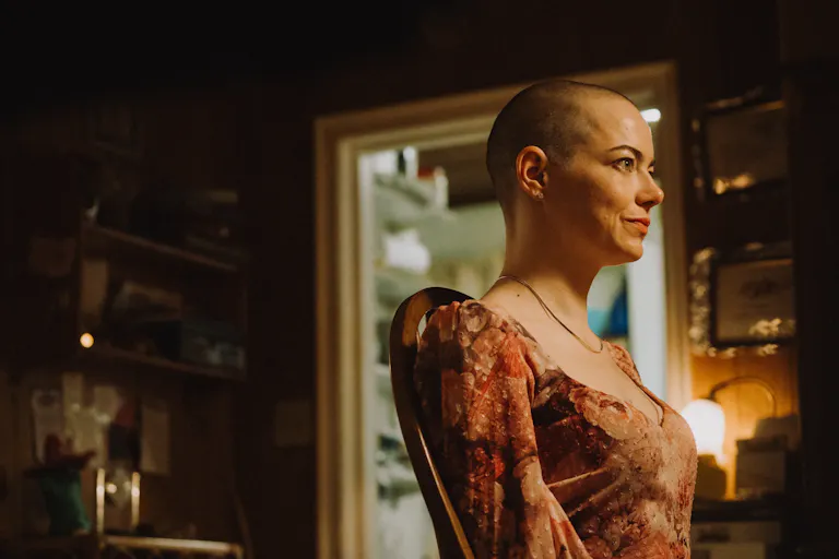 A bald, white woman sits in a chair looking to the rights while wearing an orange patterned dress.