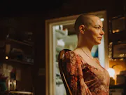 A bald, white woman sits in a chair looking to the rights while wearing an orange patterned dress.