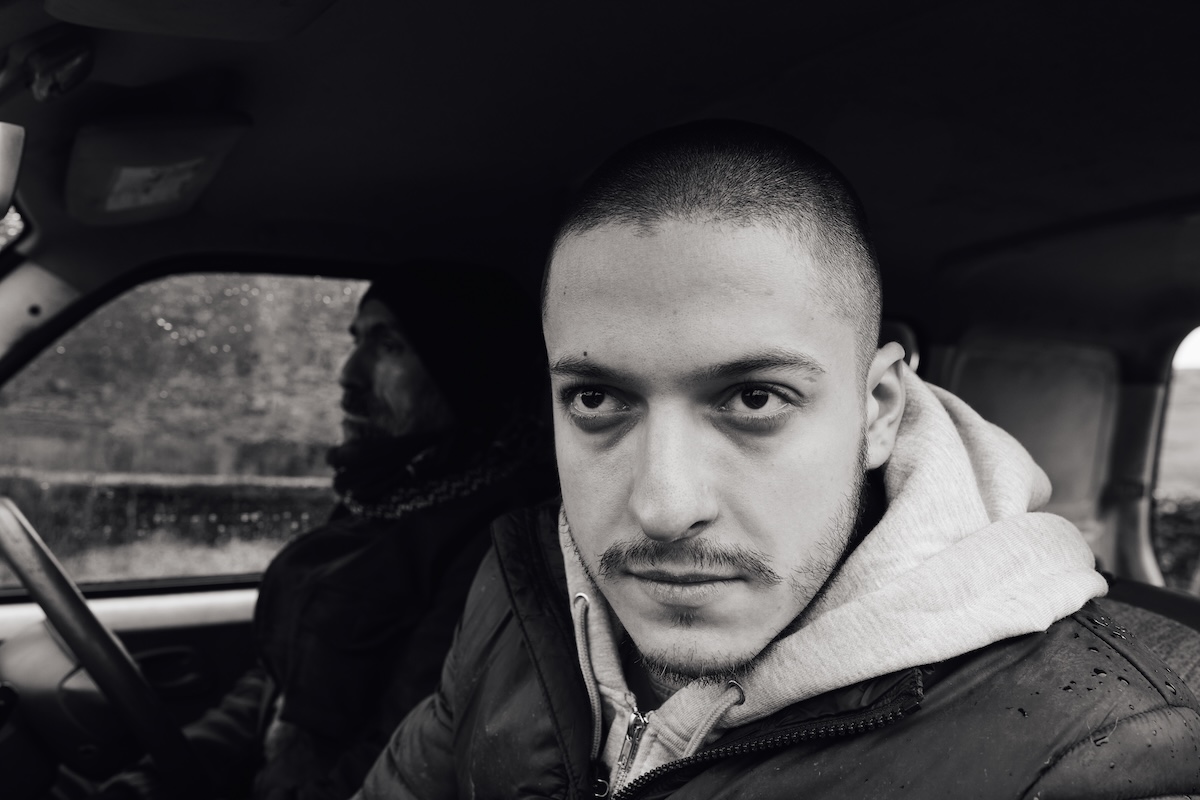 A black and white photo with one man in the foreground looking intensely out the car window and brightly lit, and one man behind him in the shadows driving the car