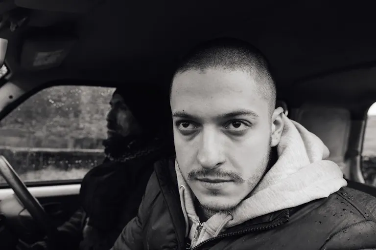 A black and white photo with one man in the foreground looking intensely out the car window and brightly lit, and one man behind him in the shadows driving the car