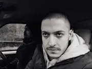 A black and white photo with one man in the foreground looking intensely out the car window and brightly lit, and one man behind him in the shadows driving the car