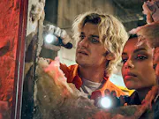 A man and a woman look questioningly through a hole in the wall while shining flashlights through it.