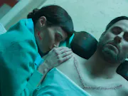 A woman lies over aa man with a v shaped scar across his chest, she holds her hand on his shoulder with a line of blood splattered on her jaw line.