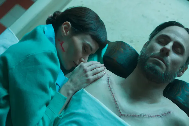 A woman lies over aa man with a v shaped scar across his chest, she holds her hand on his shoulder with a line of blood splattered on her jaw line.
