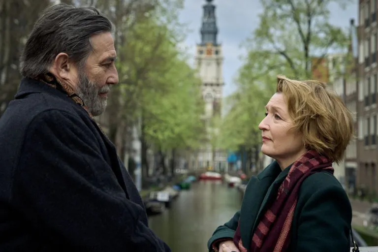 An older white man and white woman stand looking at each other infant of a canal with buildings on either side of them.