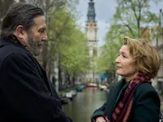 An older white man and white woman stand looking at each other infant of a canal with buildings on either side of them.