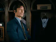 A white man in a blue suit stands with a bluetooth device in his ear in front of another suit.
