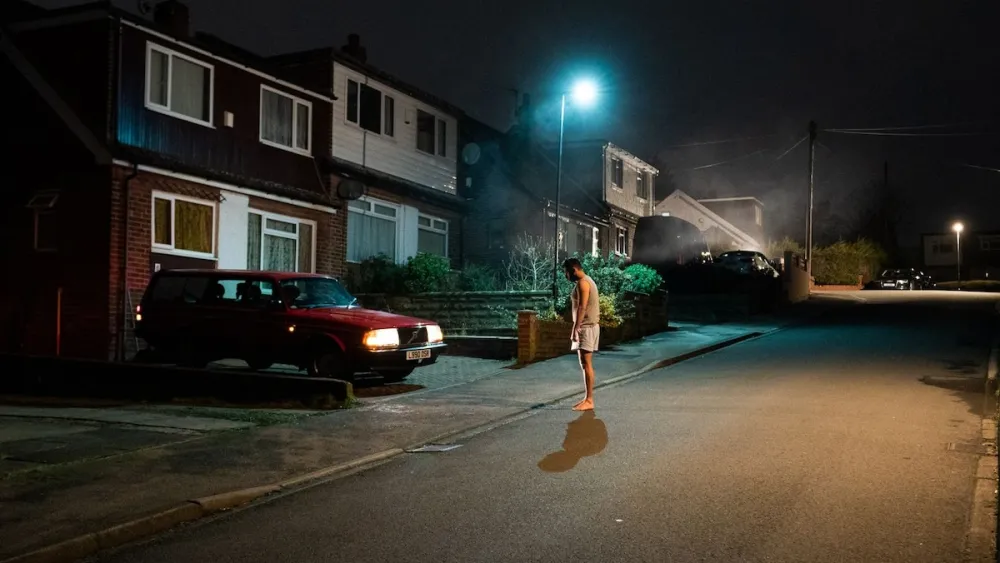 A man stands in the middle of a street at night facing a red car with its lights on.